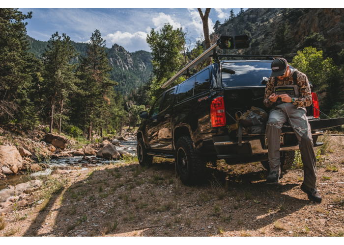 Image of a man sitting on the open bed of a black Chevy pick up truck with a matching black bed cap. There is a silver 2-banger river quiver with an open reel box mounted to the top of the truck, above the man. The man is tying a knot in some fishing line. 