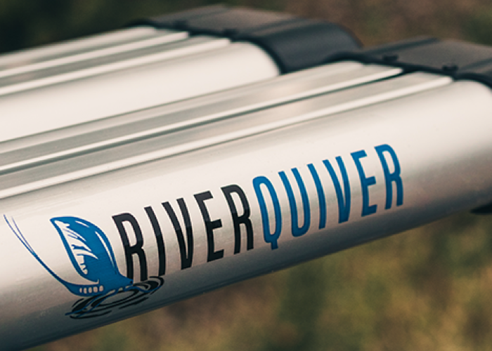 Close up of the river quiver logo on the side of the silver metal of the 4-banger river quiver’s rod casing. The logo says “river” in black text and “quiver” in blue text with a matching blue image of fishing bait hitting the water. The background is blurred out but looks vaguely like vegetation. 