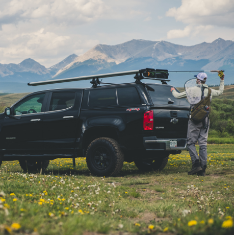 Image of a man sliding a fishing rod into a silver river quiver. The river quiver is mounted onto a black Chevy pick up truck with a matching black bed cap. The truck is parked in a beautiful field with green grass and yellow and white wild flowers. In the distance are jagged mountains with light snow covering. 